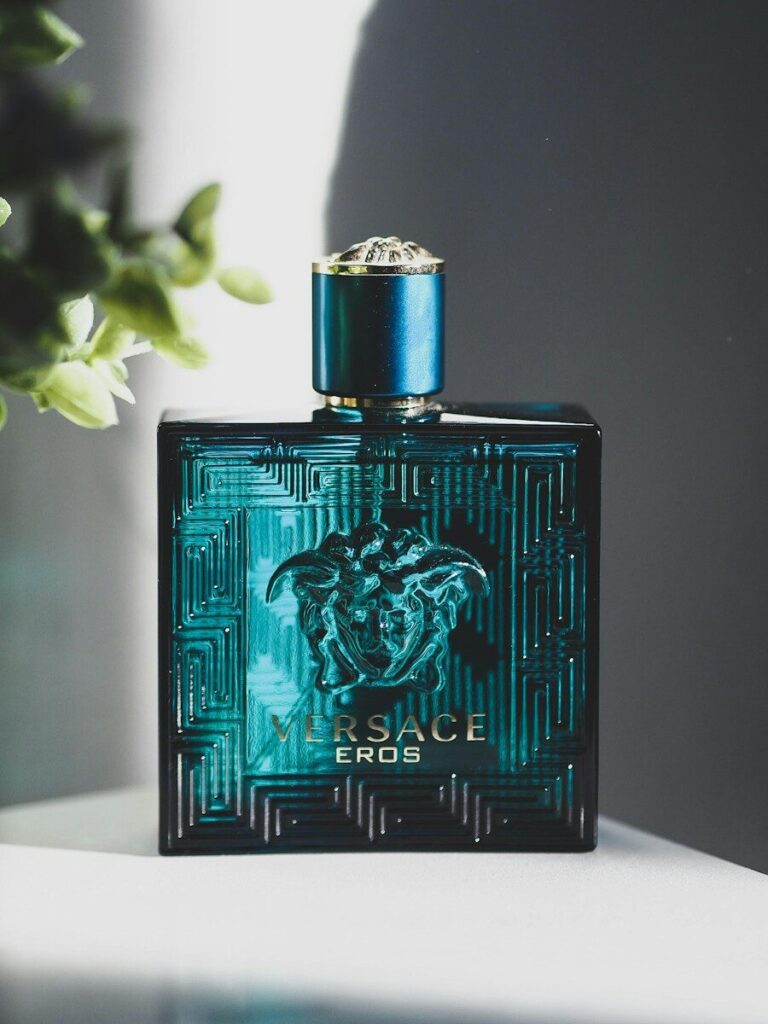 A square teal glass cologne bottle with a Medusa head emblem on a surface